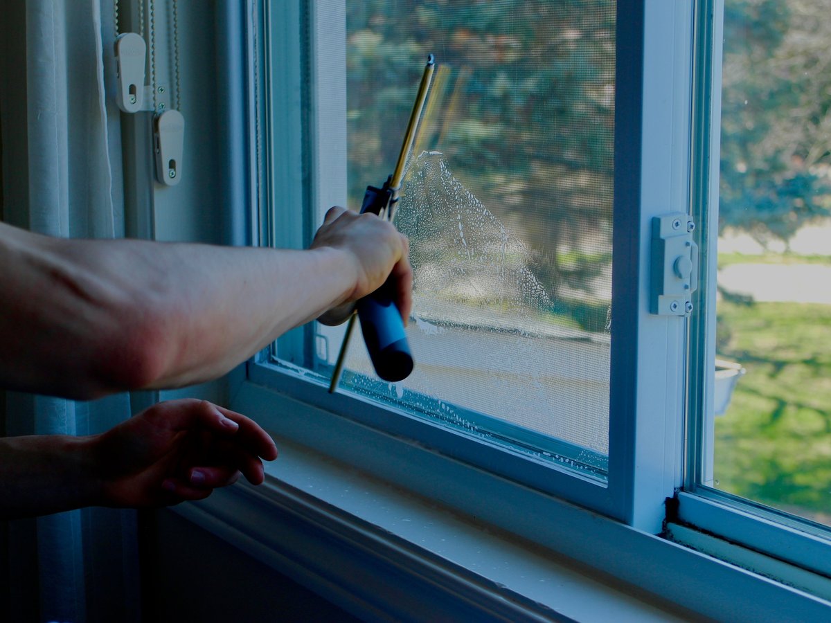 Person spraying and cleaning a blue window frame with outdoor landscape visible through the glass