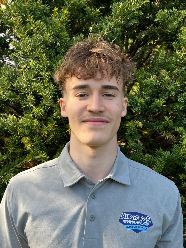 Young man with brown hair wearing a light blue AuraGlass polo shirt, smiling at camera with green foliage background