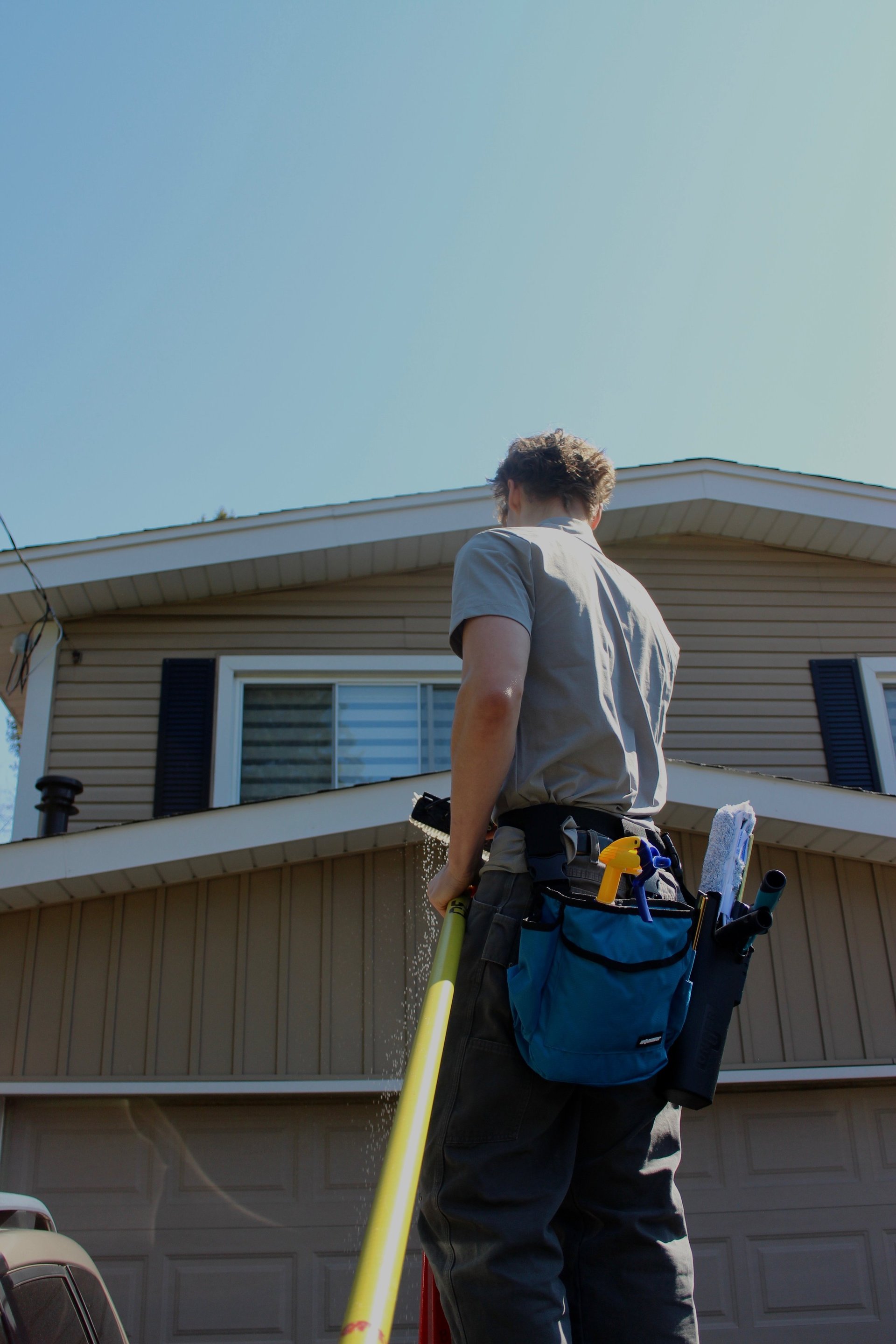 Worker on ladder wearing tool belt inspecting two-story house exterior with clear sky background