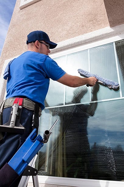 Worker in blue shirt cleaning window with squeegee using ladder against beige building